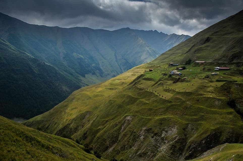 Un piccolo villaggio su un versante montuoso verde e soleggiato si affaccia su una vasta valle sotto un cielo nuvoloso.