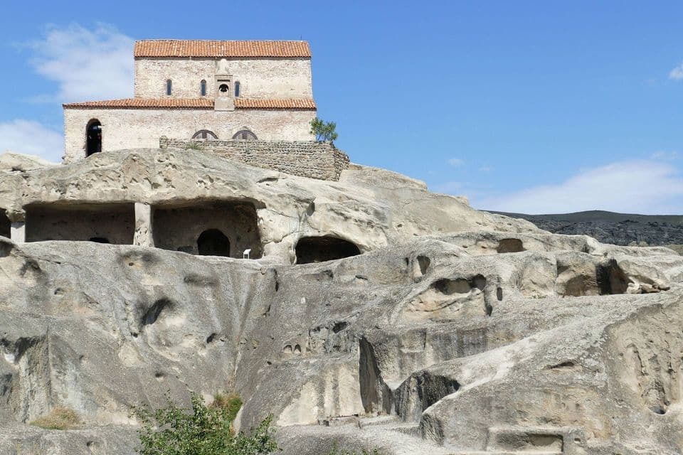 Un antico edificio in pietra con tetto in tegole rosse si erge su una grande formazione rocciosa costellata di grotte, sotto un cielo azzurro.