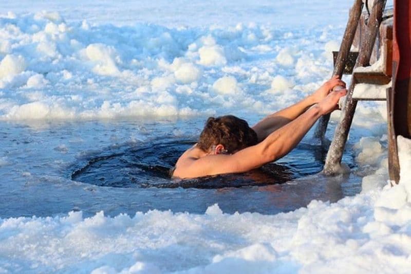 A person holds onto a wooden ladder while taking a polar plunge in a hole cut through a frozen, snow-covered body of water.
