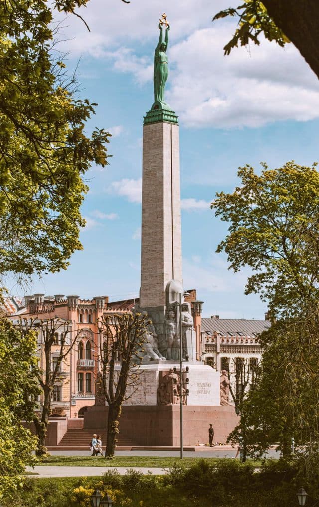 Un alto obelisco in pietra con una statua in cima, visto da un parco con alberi verdi che incorniciano il monumento sullo sfondo di un cielo blu.
