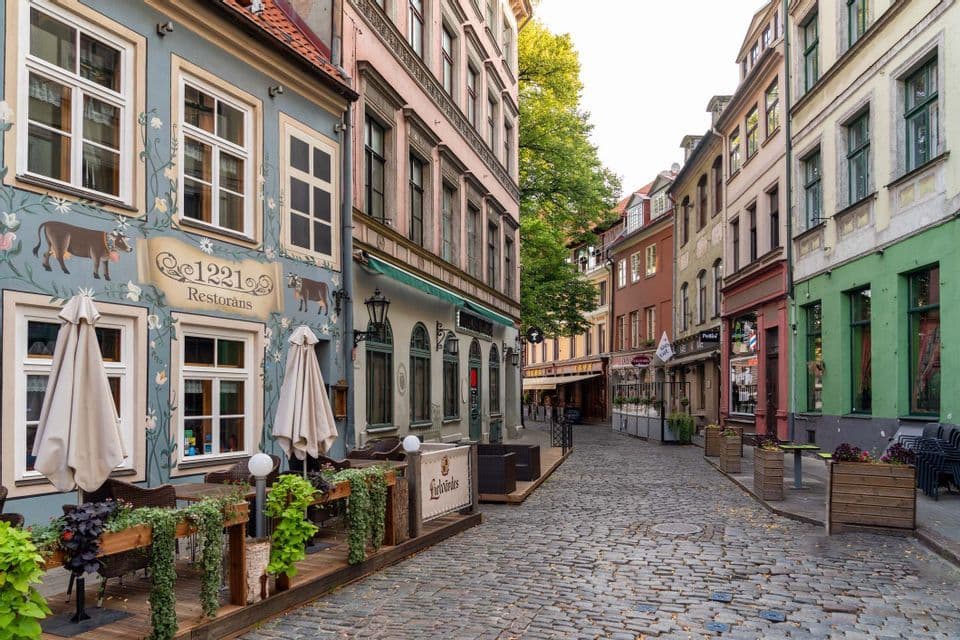 A narrow cobblestone street winds through colorful historic buildings, with an outdoor restaurant patio in the foreground.