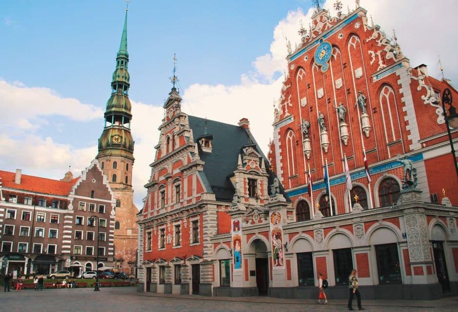 Ornate red brick buildings and a tall church spire in a historic city square under a blue sky with clouds.