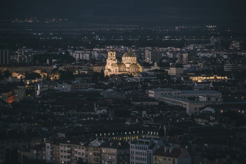 Una cattedrale illuminata con cupole dorate si staglia in un vasto panorama urbano notturno, vista da un punto elevato.