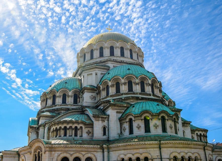 Una gran catedral con una cúpula dorada grande y cúpulas turquesa más pequeñas, vista desde abajo contra un cielo azul parcialmente nublado.