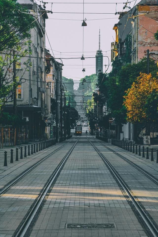 Una vista extensa de una calle urbana con vías de tranvía que se extienden entre edificios y árboles hacia un tranvía y una torre a lo lejos.