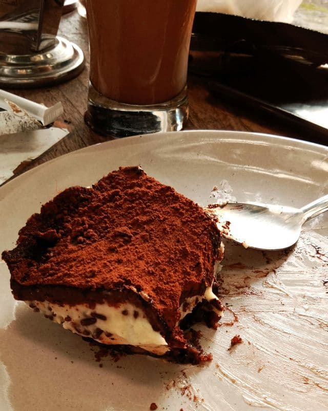 Un primo piano di una fetta di tiramisù parzialmente mangiata, spolverata di cacao, poggiata su un piatto bianco disordinato con un cucchiaino.
