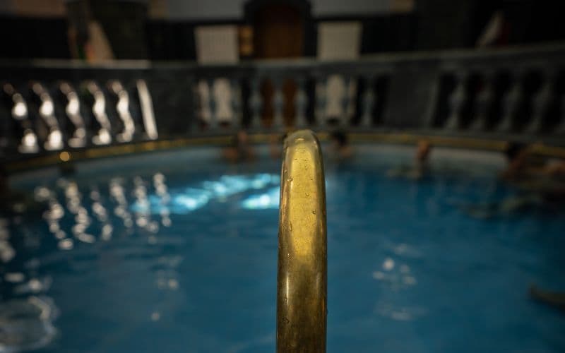 Primer plano de una barandilla dorada con vistas a un grupo de WeRoad relajándose en las aguas azules de una piscina termal cubierta.