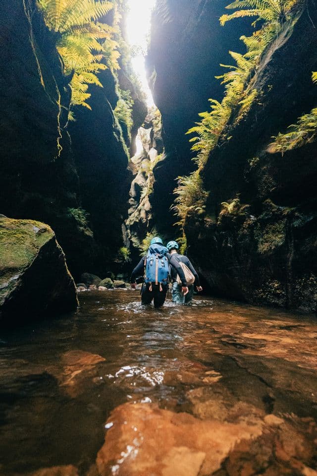 Dos personas de un viaje en grupo de WeRoad con mochilas y cascos vadeando el agua de un cañón estrecho.