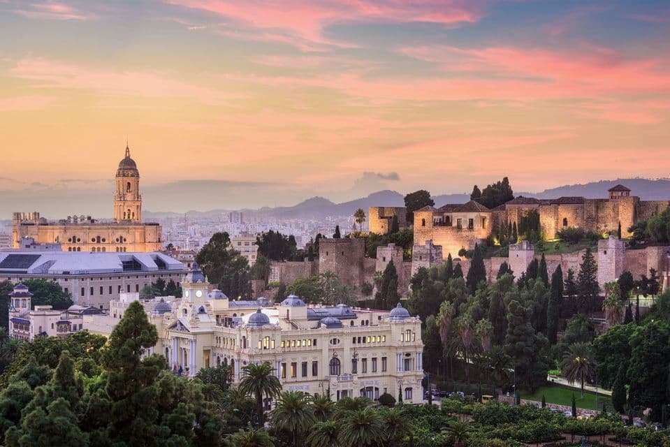 Un paisaje urbano histórico europeo con una torre de catedral y una fortaleza en la cima de una colina, visto al atardecer bajo un cielo rosa y naranja.