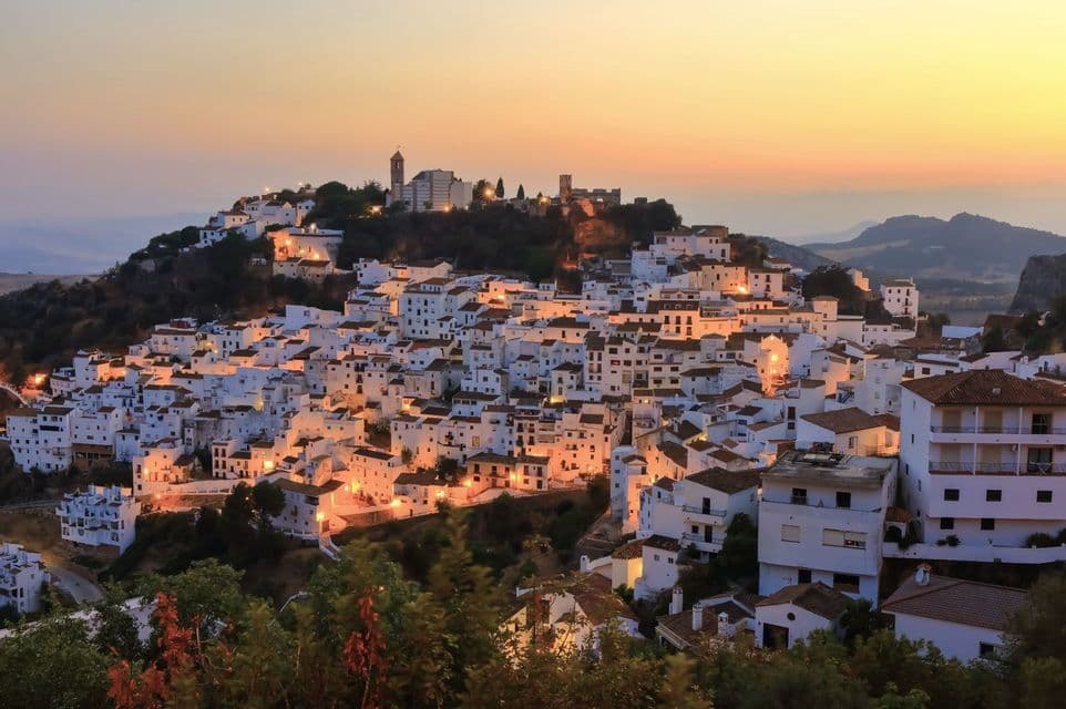Un pueblo de casas de paredes blancas apiñadas en una ladera, con luces brillando al atardecer contra un cálido cielo anaranjado.