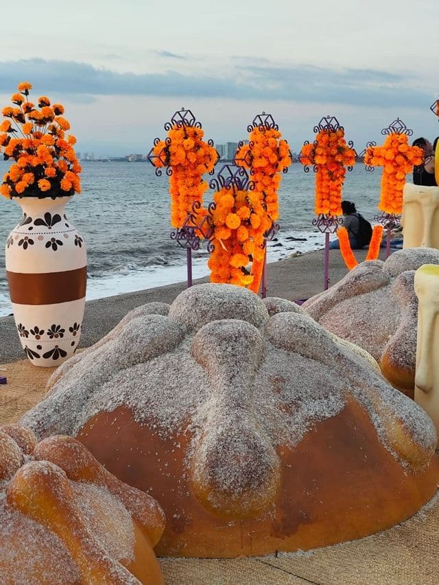 Un altare con grandi sculture di Pan de Muerto e croci di calendula esposte su una spiaggia in riva all'oceano.