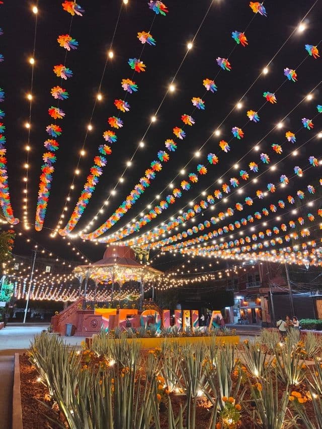 Ghirlande di luci e colorati fiori di carta addobbano una piazza cittadina di notte, con un gazebo, piante di agave e un grande cartello 'Tequila'.