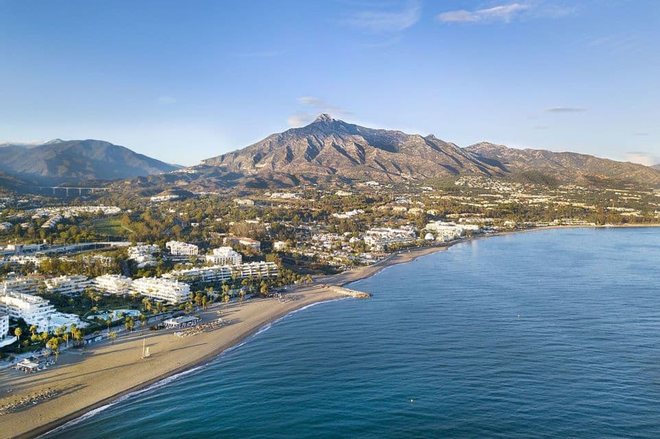 Una vista aérea de una ciudad costera con edificios blancos a lo largo de una playa de arena, con una gran montaña de fondo bajo un cielo despejado.