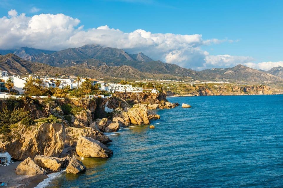 Un pueblo costero con edificios blancos se asienta en un acantilado rocoso junto al mar azul, con una cordillera al fondo bajo un cielo parcialmente nublado.
