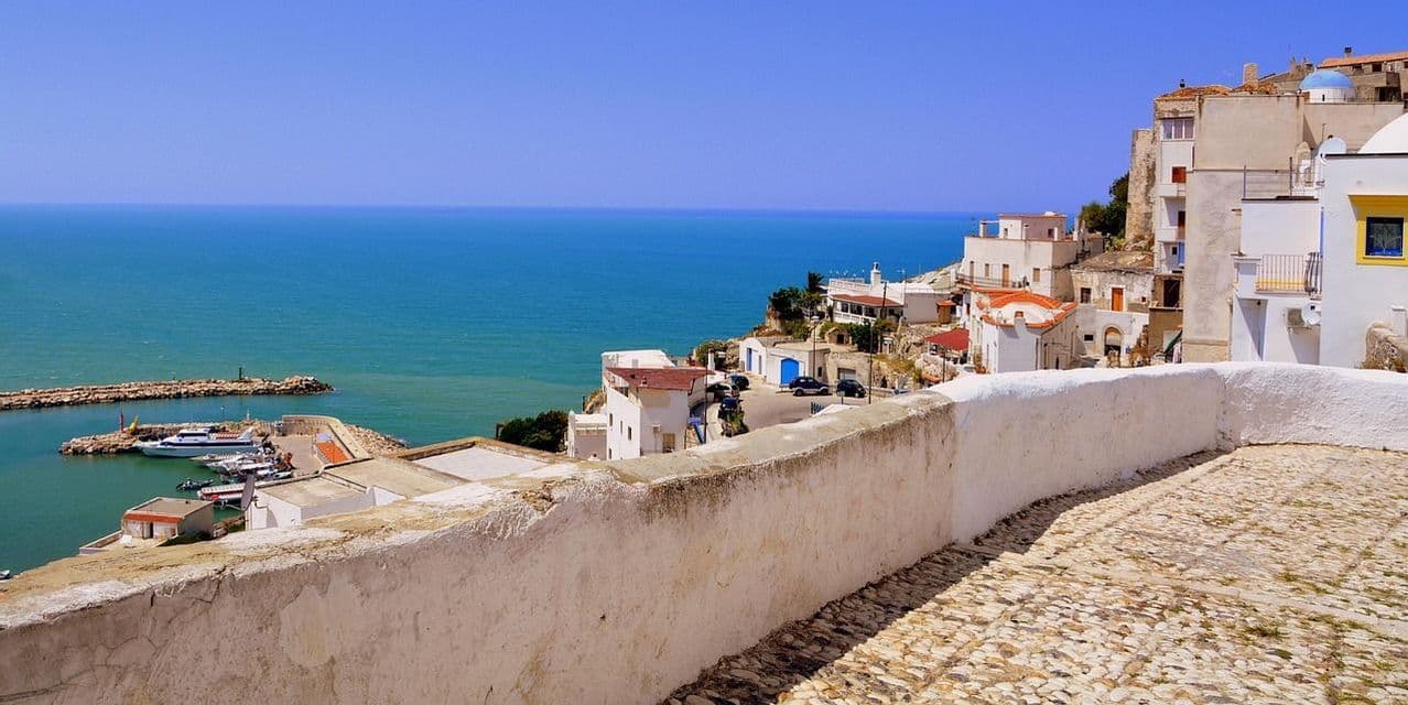 Un sentiero di ciottoli e un basso muro bianco si affacciano su una città di mare con edifici bianchi, un porto e il mare turchese sotto un cielo limpido.