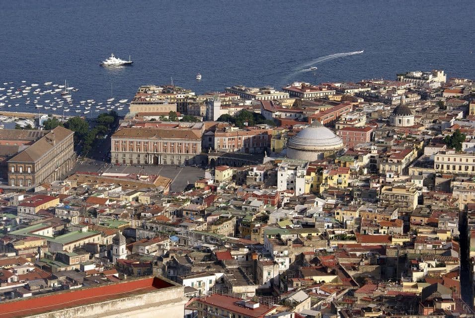 Una vista aerea di una storica città costiera con tetti in terracotta, una grande piazza e un edificio a cupola accanto al mare blu con barche.