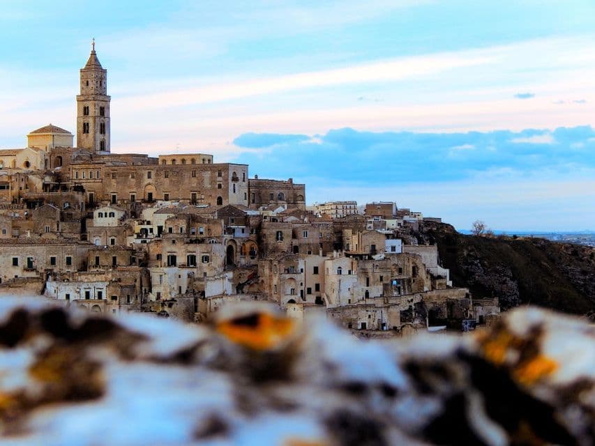 Una città collinare con antichi edifici in pietra e un campanile, sotto un cielo parzialmente nuvoloso, vista da un'angolazione bassa su un primo piano sfocato.