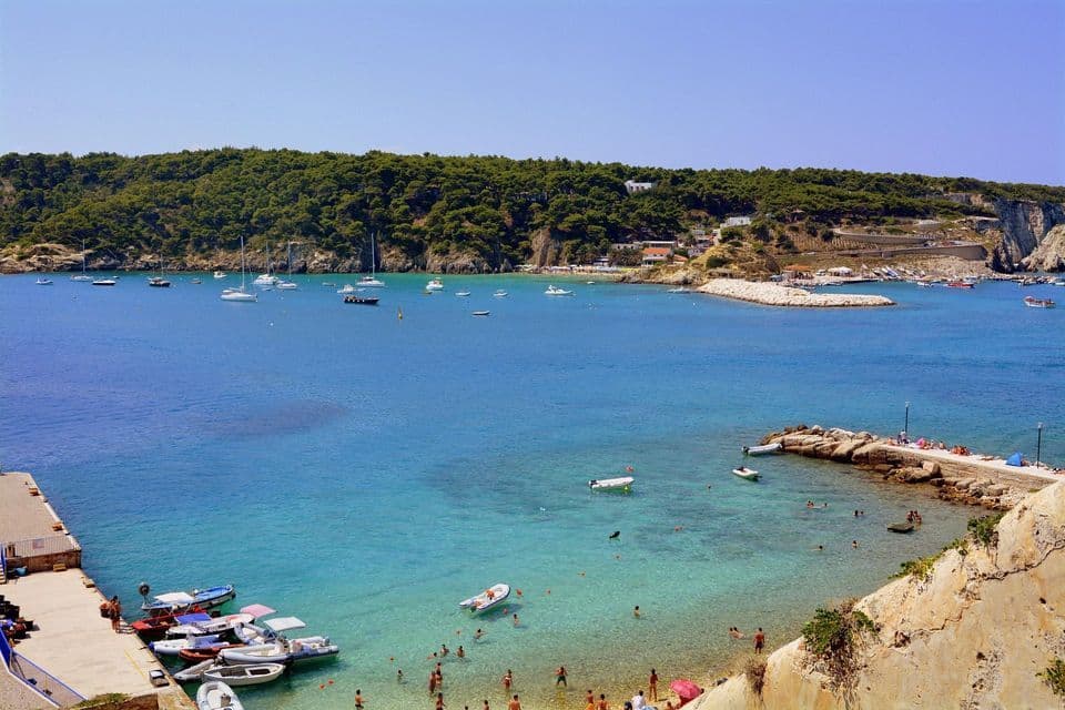 Veduta aerea di una baia costiera con acqua turchese cristallina, numerose imbarcazioni e persone che nuotano vicino ai pontili in una giornata di sole.