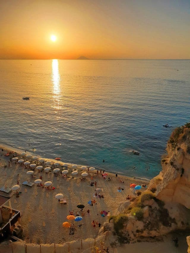 Vista dall'alto di una spiaggia sabbiosa affollata con ombrelloni colorati mentre il sole tramonta sull'oceano.