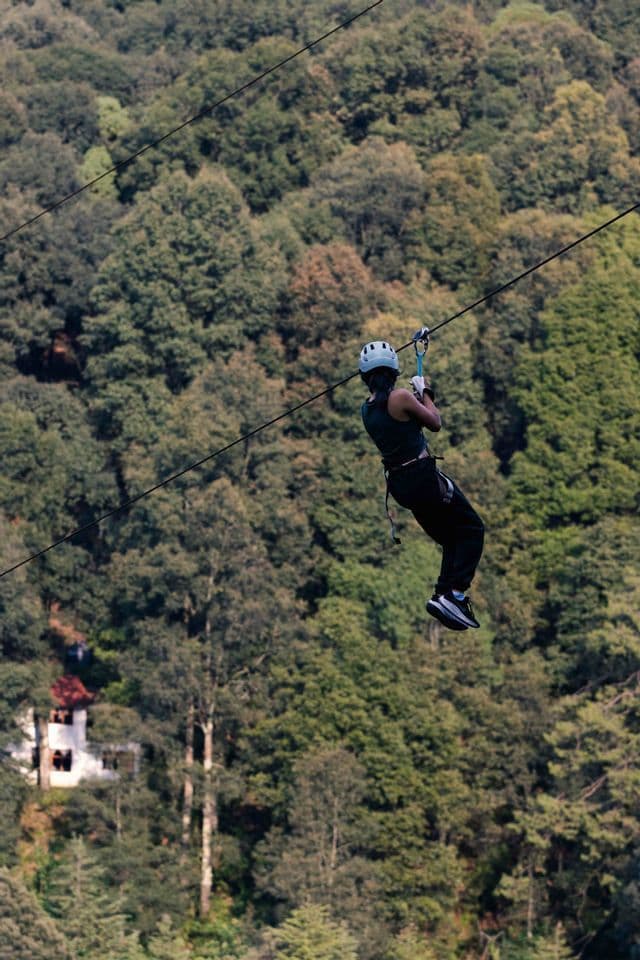 Una persona con il casco scivola su una zipline in alto sopra una fitta foresta verde che ricopre un pendio.