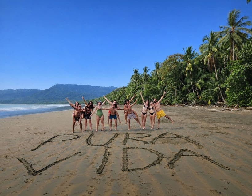 A WeRoad group trip poses on a tropical beach behind the words 'PURA VIDA' written in the sand.