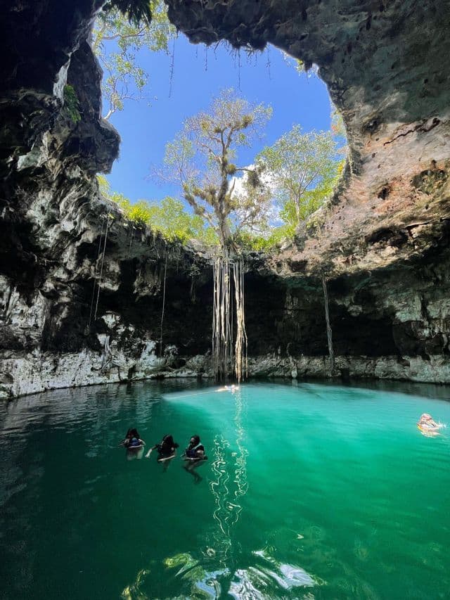 Un viaggio di gruppo WeRoad nuota nelle acque turchesi di un cenote, con la luce del sole che filtra attraverso una grande apertura nel soffitto della grotta.