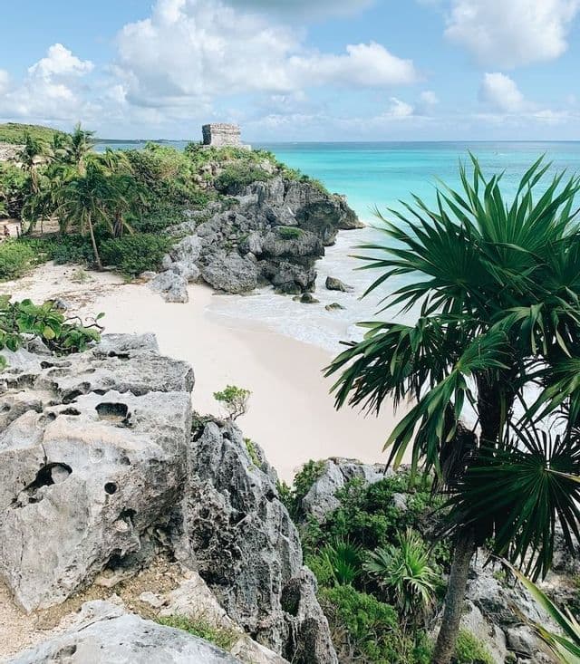 Una antigua ruina de piedra en un acantilado rocoso domina una playa de arena blanca y un mar turquesa, con hojas de palma en primer plano.