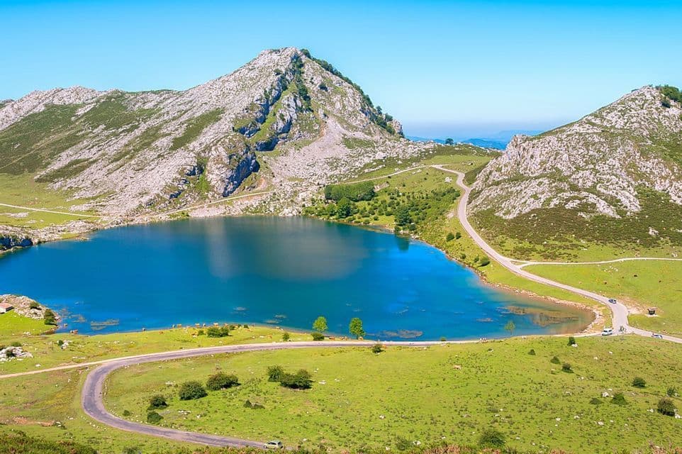 Una vista aérea de una carretera serpenteante junto a un lago azul vibrante, rodeada de colinas verdes y montañas rocosas bajo un cielo despejado.