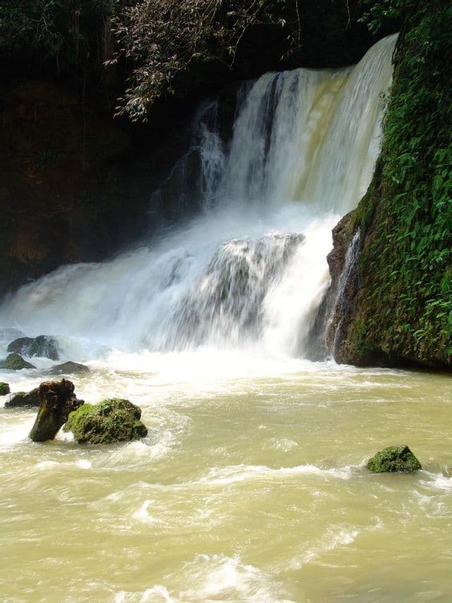 Un'ampia cascata scende da una scogliera rocciosa con vegetazione verde in un fiume agitato con pietre coperte di muschio.
