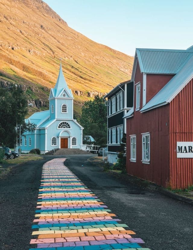 Un sentiero dipinto con i colori dell'arcobaleno su una strada asfaltata conduce a una chiesa azzurra ai piedi di una montagna illuminata dal sole.