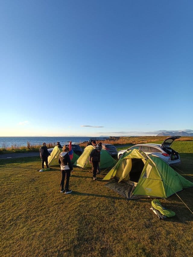 Eine WeRoad-Gruppenreise mit mehreren grünen Zelten auf einem grasbewachsenen Campingplatz direkt am Meer unter einem klaren blauen Himmel.