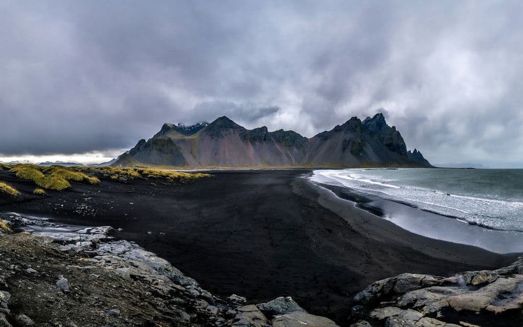 Ein Panoramablick auf einen schwarzen Sandstrand mit Meereswellen, die vor einer zerklüfteten Bergkette unter einem bewölkten Himmel auf das Ufer treffen.
