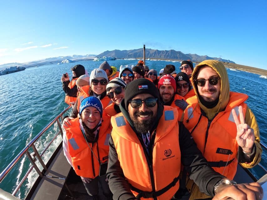 Un viaggio di gruppo WeRoad con giubbotti di salvataggio arancioni si scatta un selfie su una barca, navigando attraverso uno specchio d'acqua con iceberg e montagne.