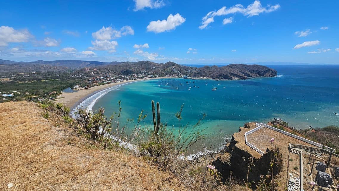 Panoramablick von einer Anhöhe auf eine türkisfarbene Bucht mit Booten, einem Sandstrand und einer Küstenstadt unter blauem Himmel.