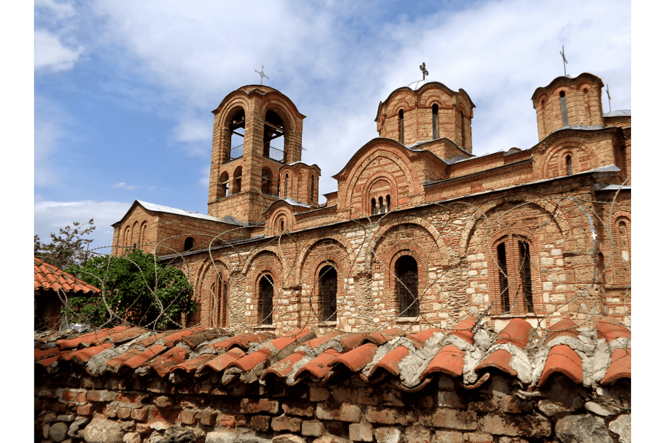 Una storica chiesa in pietra con cupole e un campanile, visibile oltre un muro con tegole e filo spinato sotto un cielo nuvoloso.