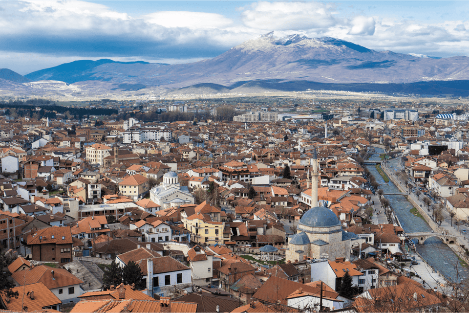 Una vista aerea di una città con edifici dai tetti in terracotta, una moschea e un fiume, con montagne innevate sullo sfondo.