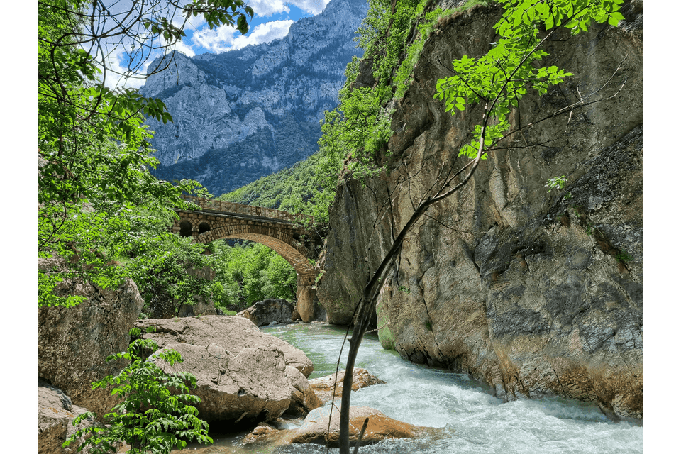 Un ponte ad arco in pietra attraversa un fiume impetuoso in una gola di montagna lussureggiante sotto un cielo parzialmente nuvoloso.