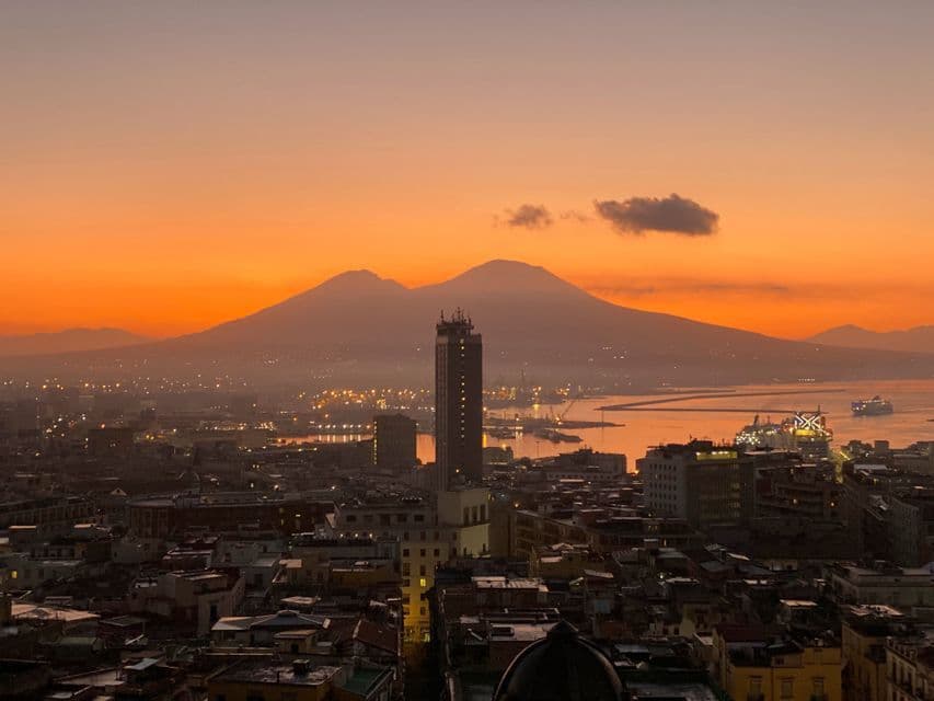 Une vue aérienne d'une ville côtière tentaculaire avec une grande montagne à deux pics en arrière-plan au coucher du soleil, sous un ciel orange éclatant.