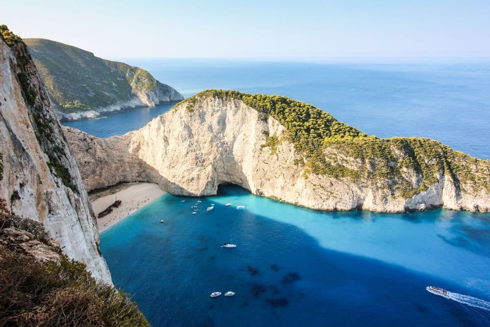 Vista aerea di una caletta isolata con un relitto su una spiaggia di sabbia bianca, circondata da scogliere e acqua turchese con barche.
