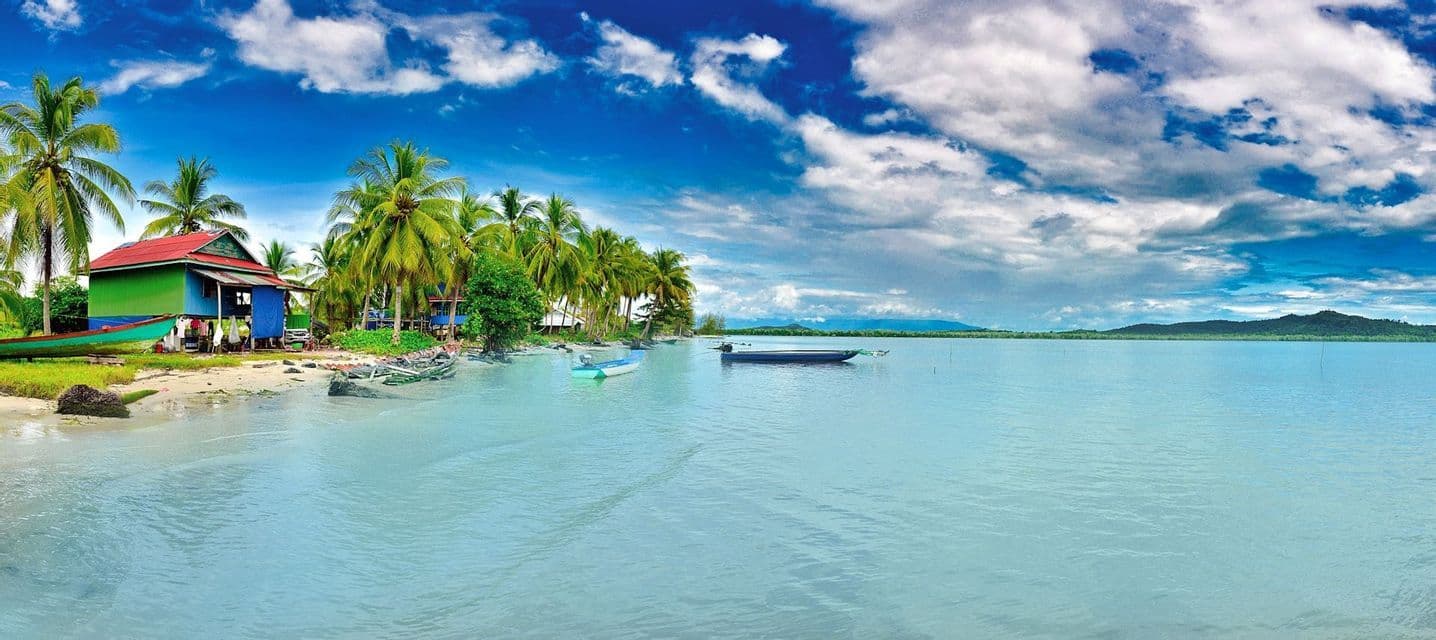 Una vista panoramica di una spiaggia tropicale con una palafitta colorata, palme e barche su acqua calma e azzurra sotto un cielo nuvoloso.