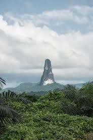 Una guglia rocciosa alta e affusolata si erge da una giungla verde e rigogliosa sotto un cielo nuvoloso.
