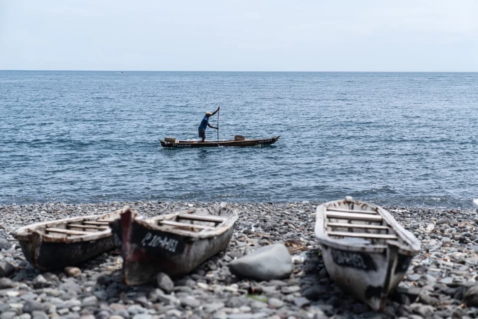 Un uomo su una barca di legno usa un palo per navigare il mare, con diverse altre barche ormeggiate sulla riva rocciosa in primo piano.