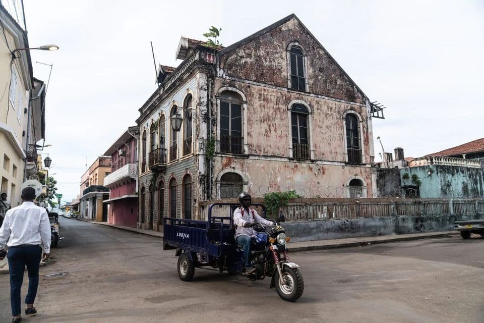 Un uomo guida un veicolo a tre ruote su una strada oltre vecchi edifici in stile coloniale e rovinati dal tempo.