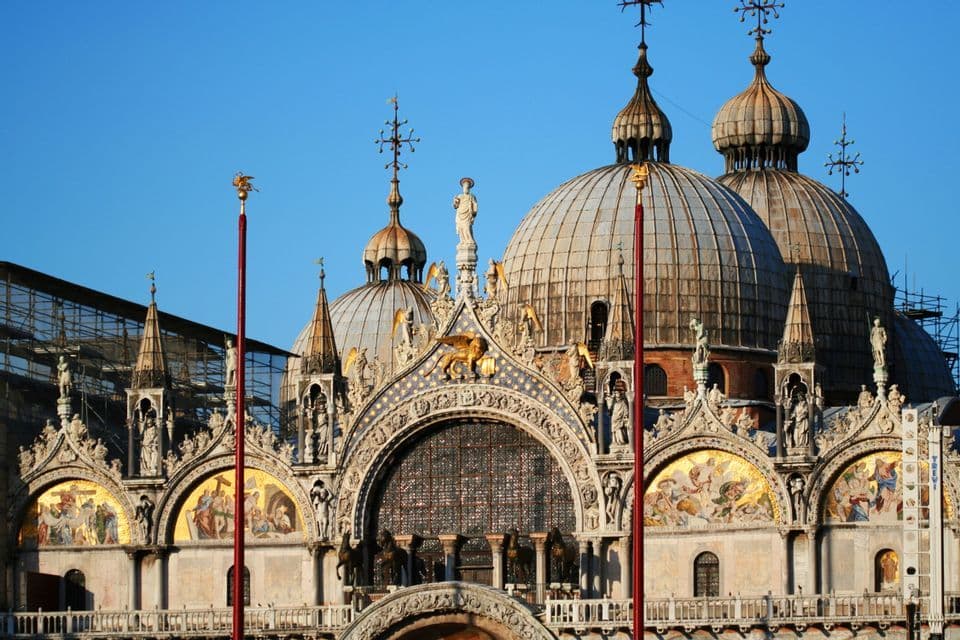 La façade supérieure ornée d'une basilique, avec de grandes coupoles, des mosaïques dorées et des sculptures détaillées, sous un ciel bleu clair.