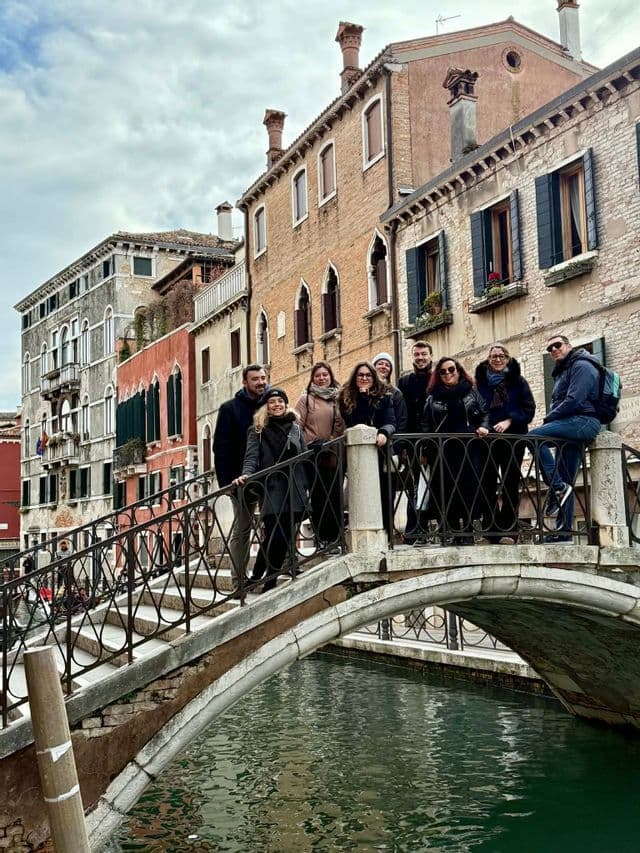 Un groupe WeRoad en voyage pose sur un pont de pierre au-dessus d'un canal, avec des bâtiments européens traditionnels en arrière-plan.