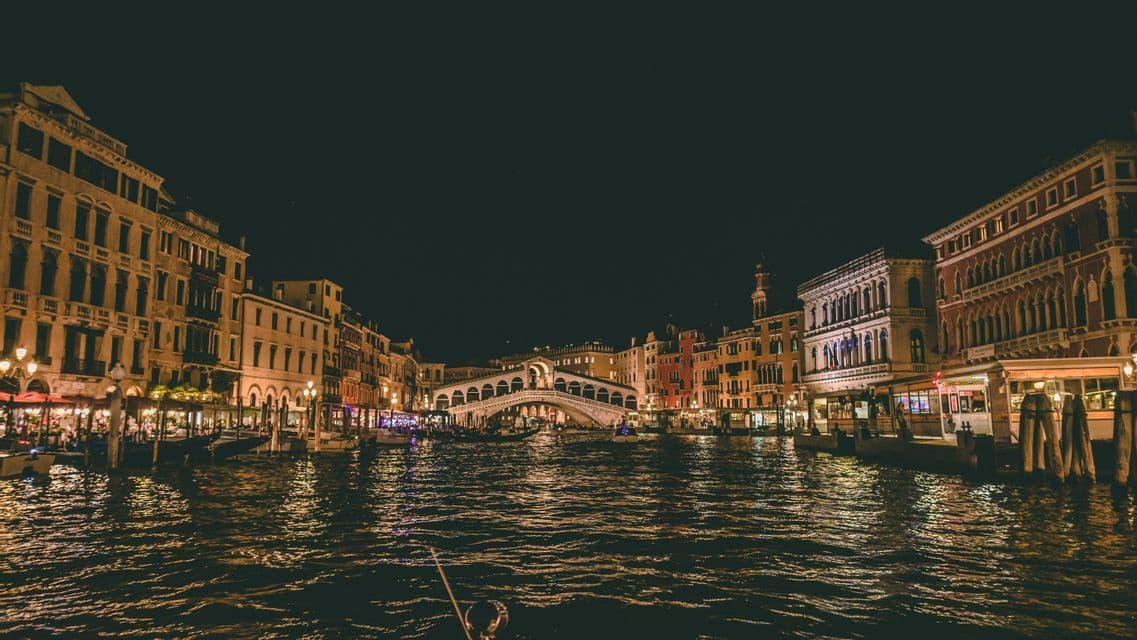 Des bâtiments illuminés bordent un large canal la nuit, avec un pont en arc de pierre en arrière-plan et des reflets sur l'eau.