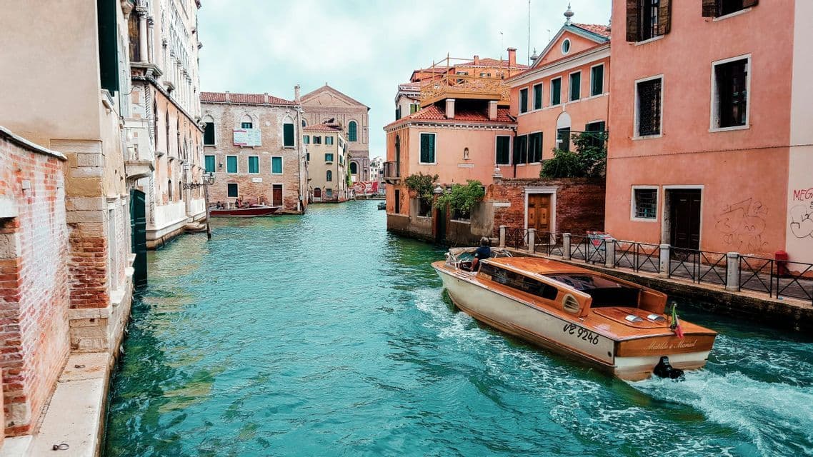 Un bateau à moteur en bois navigue sur un canal étroit bordé de bâtiments historiques et colorés, créant un sillage dans l'eau turquoise.