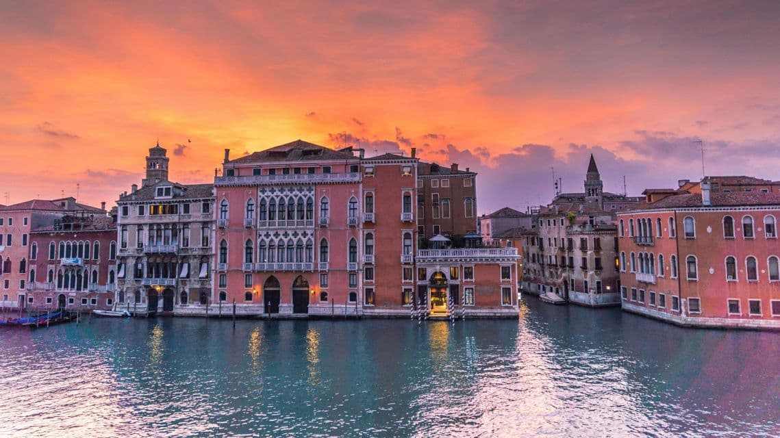 De magnifiques bâtiments historiques bordent un large canal, illuminés par un coucher de soleil vibrant aux teintes orange et violet.