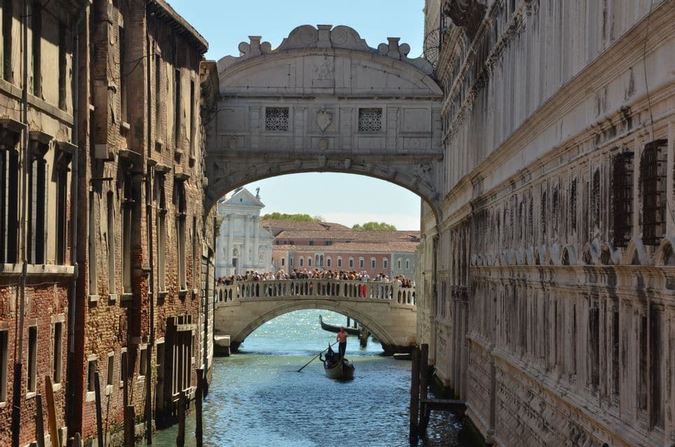 Un pont de pierre couvert enjambe un étroit canal, sous lequel passe une gondole, avec des touristes sur un autre pont en arrière-plan.