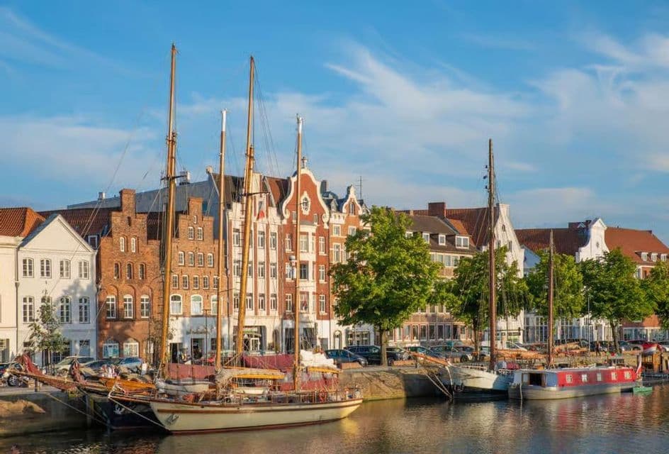 Barche a vela ormeggiate in un canale di fronte a una fila di edifici tradizionali in mattoni sotto un cielo azzurro e limpido.
